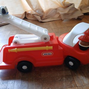May include: A red plastic toy fire engine featuring a white extendable ladder, a yellow hose attachment on the side, and a white driver cabin. The Little Tikes logo is printed on the side, and two small figures wearing red helmets are seated inside.