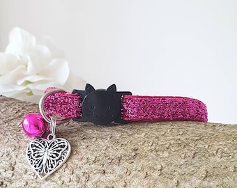May include: A pink glitter cat collar with a black cat-shaped buckle. The collar has a silver heart-shaped charm with a butterfly design and a pink bell. The collar is displayed on a piece of wood.