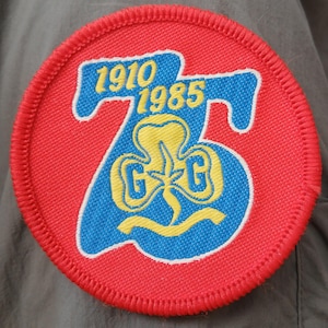 Peut inclure: Un écusson brodé circulaire avec un fond rouge et une bordure cousue rouge. Il présente un grand nombre 75 bleu avec le texte jaune 1910 et 1985 au-dessus. Au centre se trouve un emblème trèfle jaune avec les lettres G et G de chaque côté.