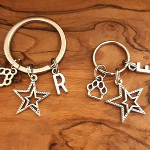 Puede incluir: Dos llaveros plateados con dijes. Un llavero tiene una huella de pata, una estrella y la letra "R". El otro llavero tiene una huella de pata, una estrella y la letra "E".