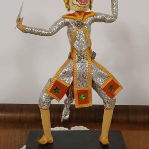 May include: A detailed figurine of a traditional Thai dancer in a dynamic pose. The figure is adorned in silver and gold attire, with a detailed mask and a small blade in one hand. It stands on a black base.