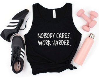 May include: Black tank top with the text "Nobody Cares. Work Harder." on it. The tank top is laying on a white surface with a pair of black sneakers, a pink water bottle, and two pink dumbbells.