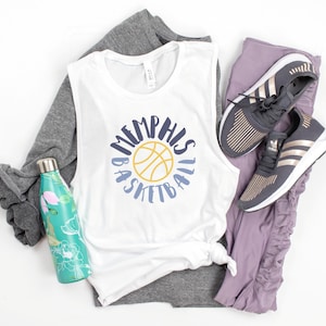 May include: White sleeveless shirt with a blue and yellow basketball graphic and the text "Memphis Basketball". A gray long-sleeved shirt, purple leggings, and a pair of black and gold sneakers are also pictured.