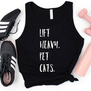 May include: Black tank top with the text "Lift Heavy. Pet Cats." on it. The tank top is laying on a white surface with a pair of black sneakers, pink dumbbells, and a pink water bottle.