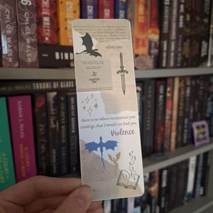 May include: A bookmark featuring a dragon, sword, and book design. The bookmark is beige with a quote that reads "there is no where in existence you could go, that I would not find you, Violence".
