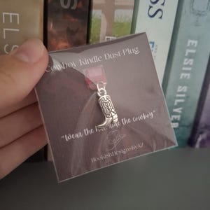 May include: A silver cowboy boot charm, designed as a Kindle dust plug, on a brown card. The card reads "Cowboy Kindle Dust Plug" and "Wear the how hige the cowboy". The product is from BookishDesignsByAZ.