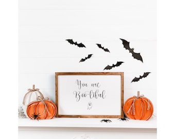May include: A white wooden sign with the text "You are Boo-tiful" in black cursive lettering. A small ghost illustration is below the text. The sign is on a white shelf with two orange and white pumpkins and black spiders. Black bats are on the white wall behind the shelf.