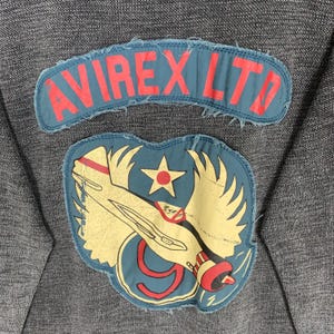 Puede incluir: Una sudadera gris oscuro con un parche azul y rojo que dice "AVIREX LTD" y un parche azul y amarillo con un avi&oacute;n de dibujos animados y una estrella.