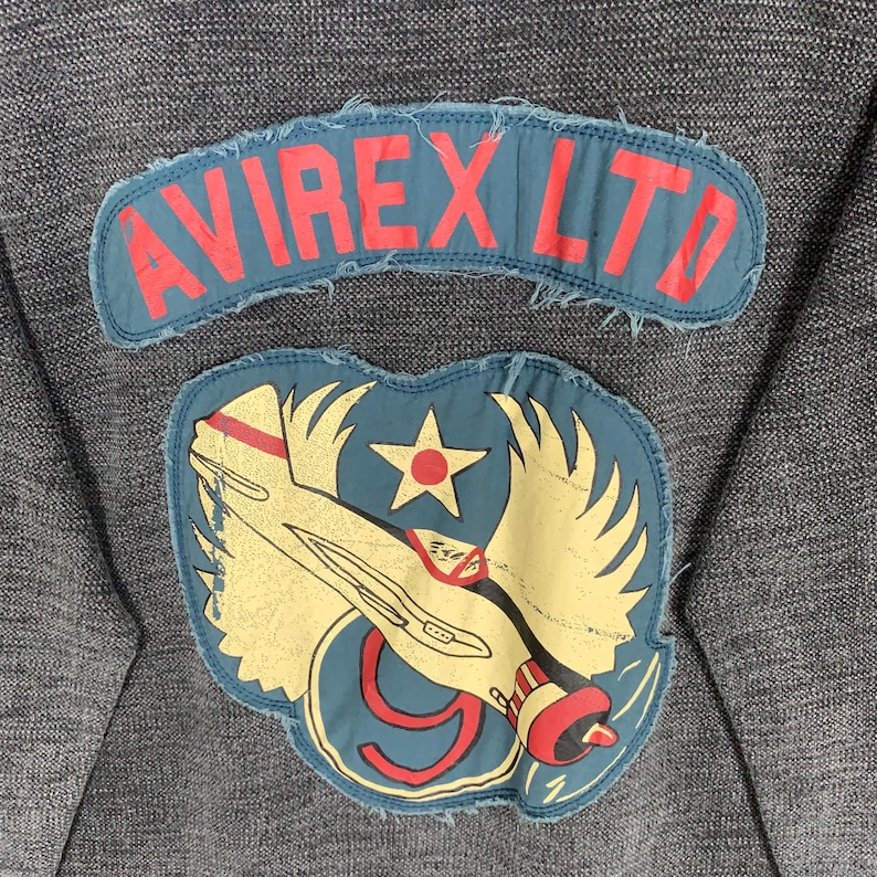 Puede incluir: Una sudadera gris oscuro con un parche azul y rojo que dice "AVIREX LTD" y un parche azul y amarillo con un avi&oacute;n de dibujos animados y una estrella.