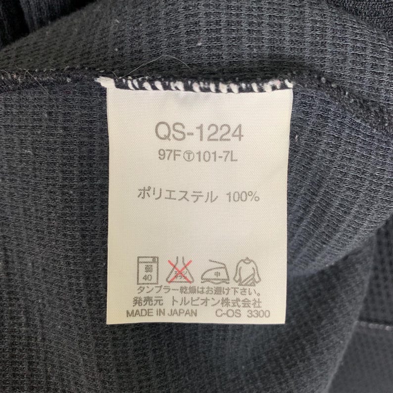 Puede incluir: Una etiqueta blanca con texto negro sobre un fondo de tela negro. La etiqueta dice "QS-1224", "97F 101-7L", "Poli&eacute;ster 100%", "Secadora a baja temperatura", "Hecho en Jap&oacute;n", "C-OS 3300", y "トルピオン株式会社".