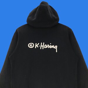 Puede incluir: Sudadera con capucha negra con un logotipo blanco que dice "&copy; K. Haring" en la espalda.