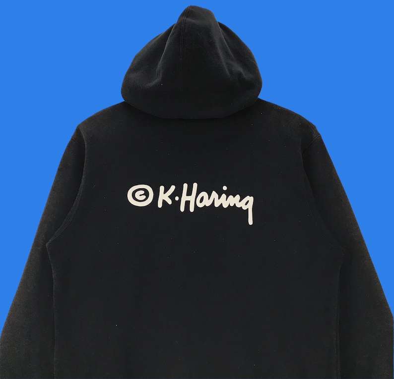 Puede incluir: Sudadera con capucha negra con un logotipo blanco que dice "&copy; K. Haring" en la espalda.