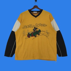 May include: Yellow, black, and white long-sleeved sweatshirt with a cartoon bear driving a green car. The sweatshirt has the text "FIORLI*SCONE*" printed on the front.