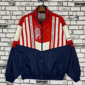 Puede incluir: Una chaqueta a rayas rojas, blancas y azules con cierre de cremallera. La chaqueta tiene un gran gráfico en la parte delantera con el texto "The World's Fastest Fighter" repetido varias veces. La chaqueta también tiene el texto "J.M. 50" en la manga derecha.