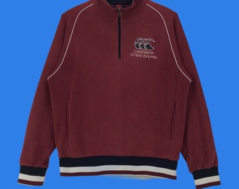 Puede incluir: Una sudadera color granate con una cremallera blanca y ribetes blancos. La sudadera tiene una banda de rayas blancas y negras en la parte inferior y en los puños. La parte delantera de la sudadera tiene un logotipo bordado en blanco que dice "Canterbury of New Zealand" con un diseño de balón de rugby.