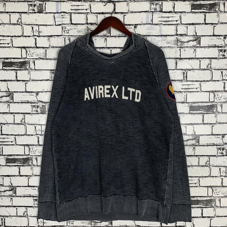 Puede incluir: Una sudadera con capucha negra de aspecto desgastado. La sudadera tiene un logotipo bordado en blanco que dice "AVIREX LTD" y un peque&ntilde;o parche en la manga izquierda.
