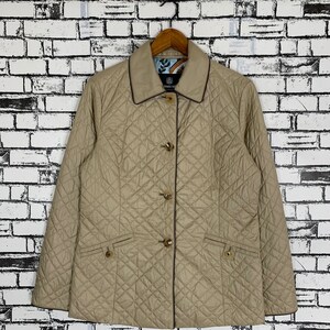 Puede incluir: Chaqueta acolchada beige con un patrón de diamantes, cierre de botones y dos bolsillos delanteros. La chaqueta tiene un ribete marrón oscuro y está colgada de una percha de madera contra una pared de ladrillos blancos.