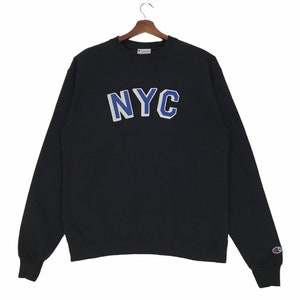 Puede incluir: Sudadera negra Champion con un logotipo NYC azul y blanco en la parte delantera. La sudadera tiene cuello redondo y mangas largas.