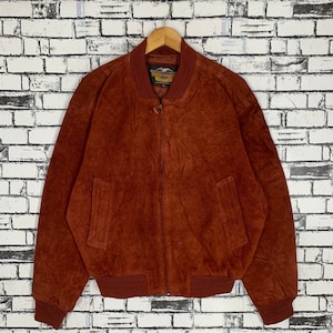 Puede incluir: Una chaqueta bomber de ante marrón con cierre de cremallera y un parche con el logotipo de Harley-Davidson en el pecho izquierdo. La chaqueta tiene cuello, puños y cintura de canalé.