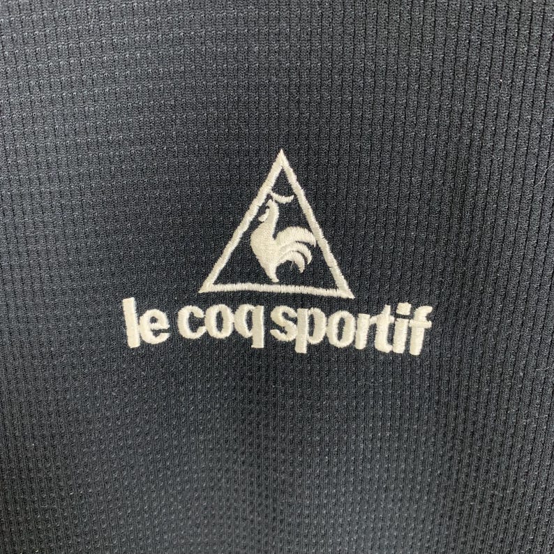 Puede incluir: Camiseta deportiva negra con un logotipo bordado blanco de un gallo en un tri&aacute;ngulo y el texto "le coq sportif".