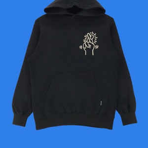 Puede incluir: Sudadera con capucha negra con un gr&aacute;fico blanco de una mano con los dedos cruzados en la parte delantera. El gr&aacute;fico est&aacute; rodeado de un c&iacute;rculo blanco con un dise&ntilde;o de estrella.