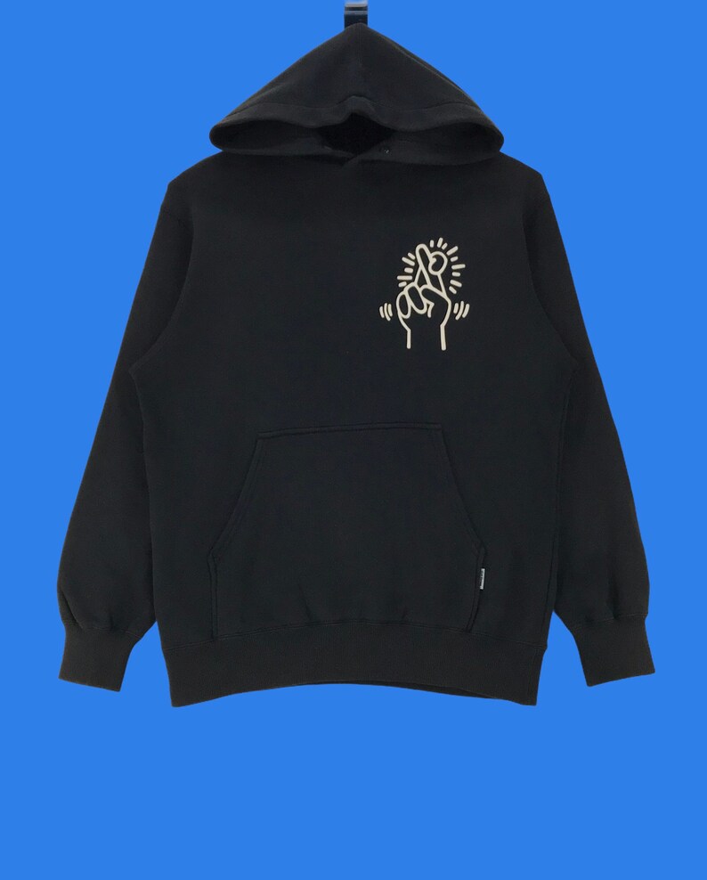 Puede incluir: Sudadera con capucha negra con un gr&aacute;fico blanco de una mano con los dedos cruzados en la parte delantera. El gr&aacute;fico est&aacute; rodeado de un c&iacute;rculo blanco con un dise&ntilde;o de estrella.