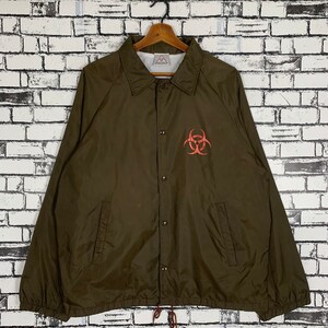 May include: A brown nylon coach jacket with a red biohazard symbol on the front. The jacket has a button closure and two pockets.
