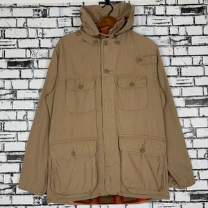 Puede incluir: Una chaqueta de color marrón claro con capucha y forro de cuadros. La chaqueta tiene cierre de botones, dos bolsillos grandes en la parte delantera y dos bolsillos más pequeños en el pecho.