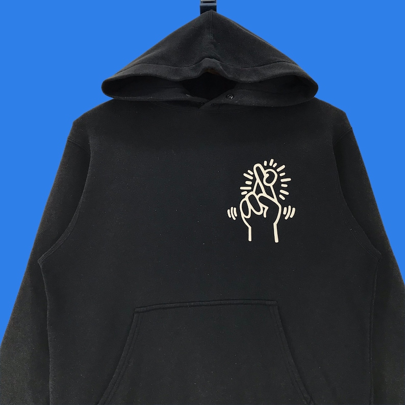 Puede incluir: Sudadera con capucha negra con un gr&aacute;fico blanco de una mano con los dedos cruzados y un dise&ntilde;o de sol radiante.