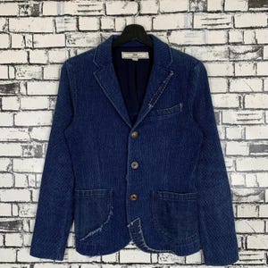 May include: A blue denim blazer with a three-button closure, two front pockets, and a distressed look. The blazer has a label that reads "Ksubi".