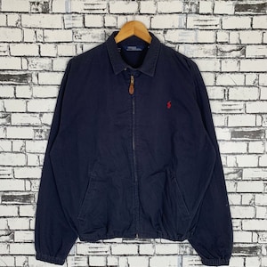 Puede incluir: Una chaqueta azul marino con cremallera con un logotipo de jugador de polo rojo en el pecho. La chaqueta tiene una cremallera marrón y una lengüeta de cuero marrón.