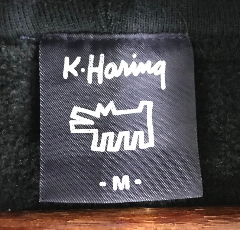 Puede incluir: Etiqueta negra con texto blanco que dice "K. Haring" y una ilustraci&oacute;n estilizada de un perro blanco. La etiqueta tambi&eacute;n tiene el texto "- M -" en blanco.