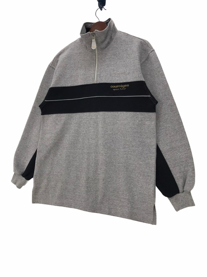 May include: A gray and black long-sleeve sweatshirt with a half-zip closure. The sweatshirt has a black stripe across the chest with the text "Courreges Sport Futur" in gold lettering.