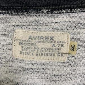 Puede incluir: Una etiqueta blanca con texto negro que dice "AVIREX STANDARD COMFORTABLE WEAR MODEL A-T5 STOCK NO. A 0063.242 GHSTRACT # 00152-ALARX-51 AVIREX CLOTHING CO." y una etiqueta blanca m&aacute;s peque&ntilde;a con texto negro que dice "2XL".