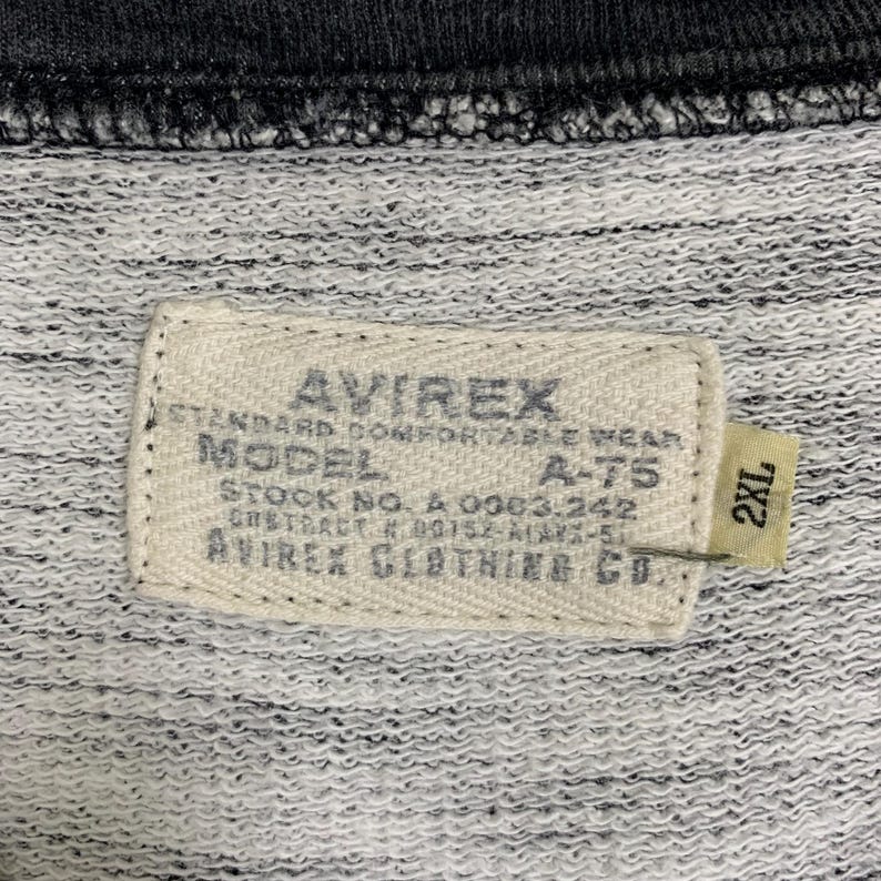 Puede incluir: Una etiqueta blanca con texto negro que dice "AVIREX STANDARD COMFORTABLE WEAR MODEL A-T5 STOCK NO. A 0063.242 GHSTRACT # 00152-ALARX-51 AVIREX CLOTHING CO." y una etiqueta blanca m&aacute;s peque&ntilde;a con texto negro que dice "2XL".