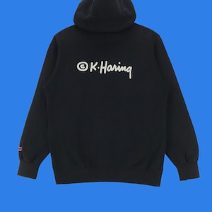 Puede incluir: Una sudadera con capucha negra con un logotipo bordado en blanco que dice "&copy;K. Haring" en la espalda.