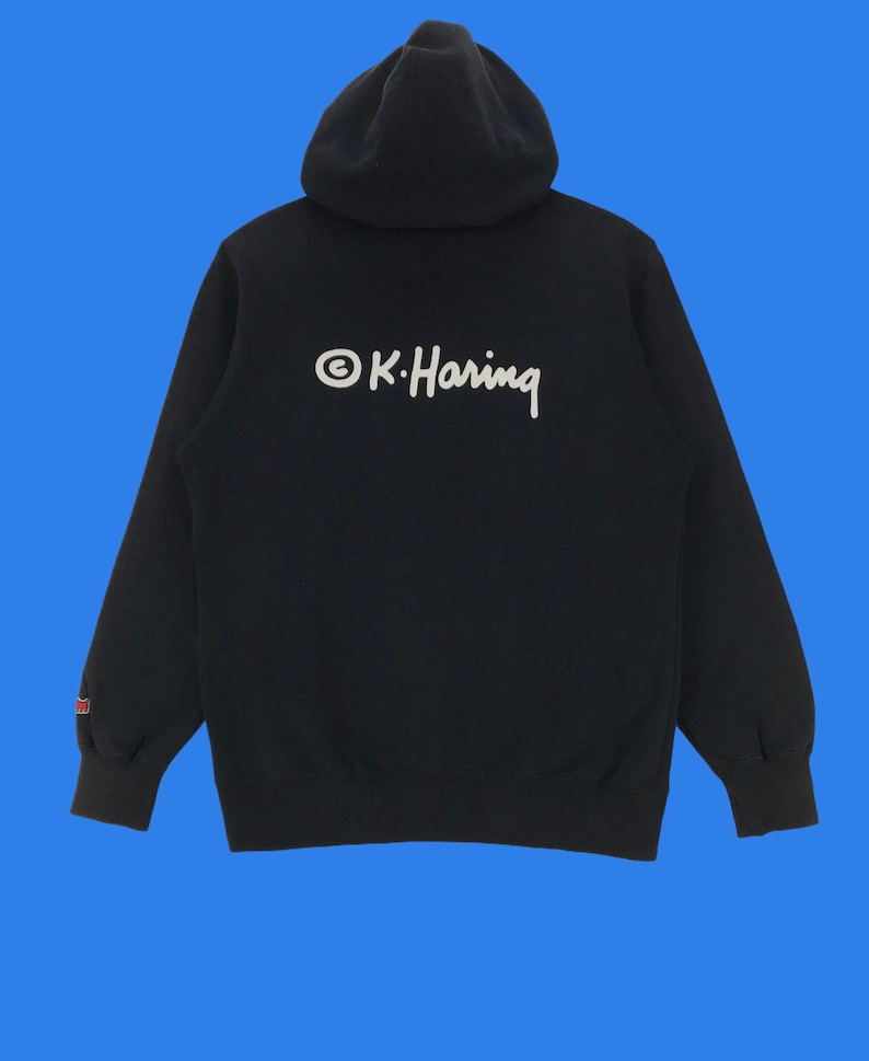 Puede incluir: Una sudadera con capucha negra con un logotipo bordado en blanco que dice "&copy;K. Haring" en la espalda.