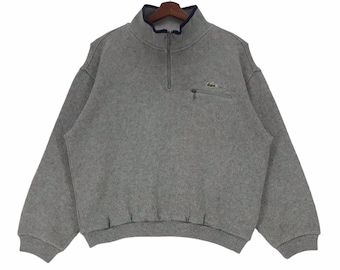 May include: A gray half-zip sweatshirt with a small green crocodile logo on the left chest and a zippered pocket on the right side.