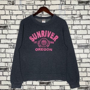 May include: Dark gray sweatshirt with the text "SUNRIVER OREGON" in pink, arched across the top. Below the text is a circular emblem with the text "EST. 1958". The sweatshirt is hanging on a wooden hanger.