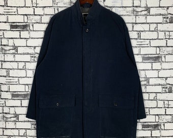 Può includere: Un cappotto blu navy con collo alto e due ampie tasche frontali. Il cappotto ha una chiusura con bottoni e un taglio lungo e dritto. Il cappotto è appeso a una gruccia di legno contro un muro di mattoni bianchi.