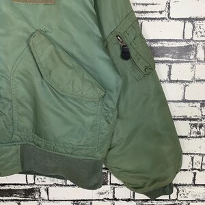 May include: A green bomber jacket with a zippered pocket on the left sleeve and a large pocket on the front. The jacket has a ribbed knit collar, cuffs, and waistband.