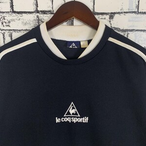 Puede incluir: Una camiseta de manga larga de punto de gofre azul marino con ribete blanco alrededor del cuello y rayas blancas en los hombros. La camiseta presenta un logotipo blanco bordado de un gallo en un tri&aacute;ngulo con el texto "le coq sportif" debajo.