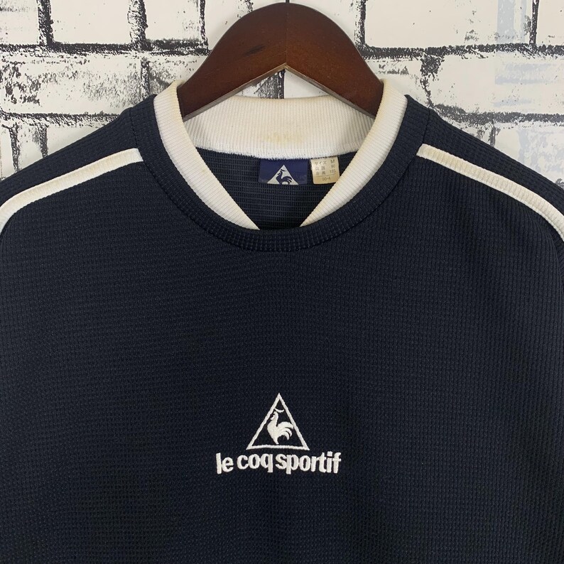 Puede incluir: Una camiseta de manga larga de punto de gofre azul marino con ribete blanco alrededor del cuello y rayas blancas en los hombros. La camiseta presenta un logotipo blanco bordado de un gallo en un tri&aacute;ngulo con el texto "le coq sportif" debajo.