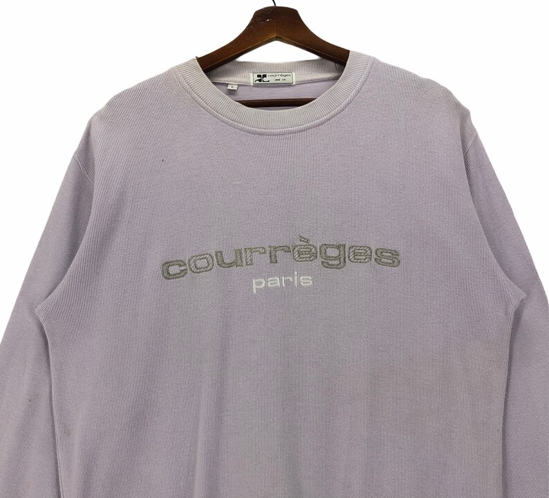 Puede incluir: Una sudadera de manga larga color lavanda con el texto "Courr&egrave;ges Paris" bordado en plata en la parte delantera.