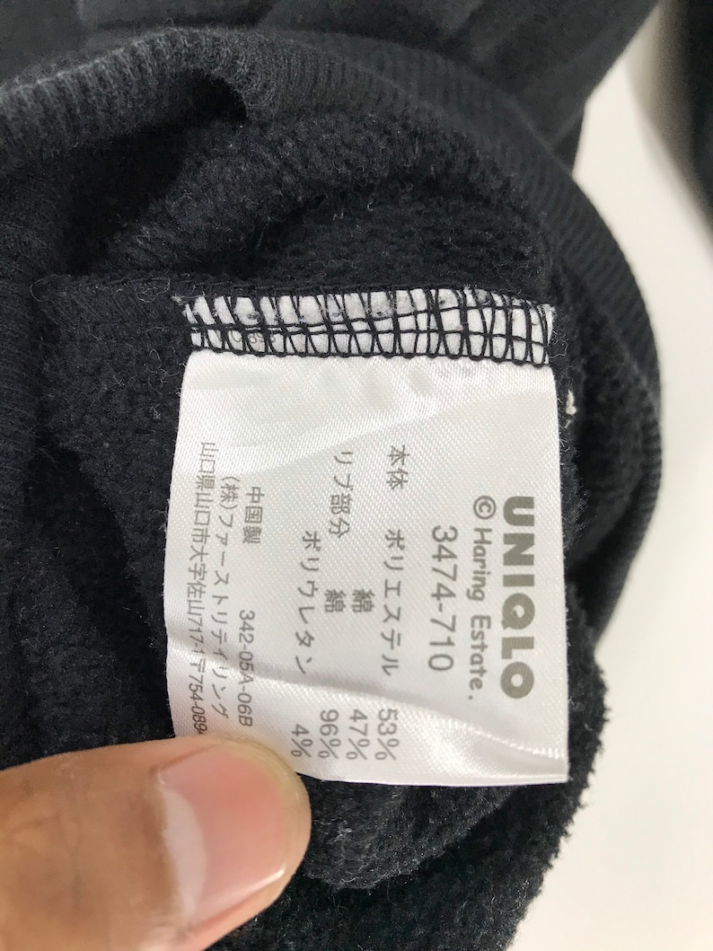 Puede incluir: Etiqueta blanca con texto negro en una sudadera negra. La etiqueta dice "UNIQLO", "Haring Estate", "3474-710", "Cuerpo: Poli&eacute;ster 53%, Algod&oacute;n 47%", "Rib: Algod&oacute;n 96%, Poliuretano 4%", "Hecho en China", "342-05A-06B", "(株)ファーストリテイリング", "Yamaguchi Prefecture, Yamaguchi City, Sayama 717-1 〒754-089".