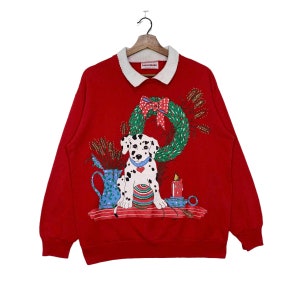 May include: Red Christmas sweatshirt with a white collar and a festive design featuring a Dalmatian puppy sitting next to a candle and a wreath. The sweatshirt has the word "remarkable" printed on the tag.