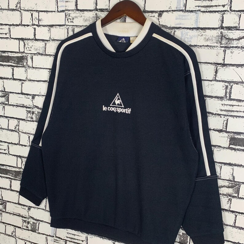 Puede incluir: Una sudadera negra de manga larga con ribetes blancos en las mangas y el cuello. La sudadera tiene un logotipo blanco bordado de un gallo en un tri&aacute;ngulo con el texto "le coq sportif".