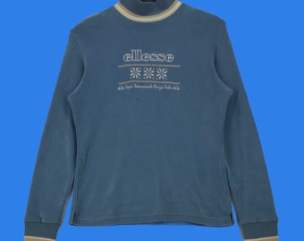 May include: A light blue long-sleeved turtleneck sweater with a white ellesse logo and a white snowflake pattern. The sweater has a white and beige trim around the neck and cuffs.