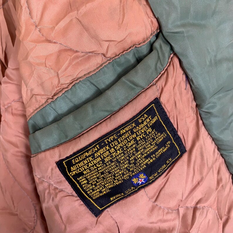 May include: A close-up of a brown and green jacket with a label that reads "EQUIPMENT - TYPE - AVIREX USA AUTHENTIC AVIREX LTD FLIGHT GARMENT SPECIFICATION FOR HS SPEED LOW LEVEL FLIGHT NO:3462 JRC35AC 2 SMC NYC PB".