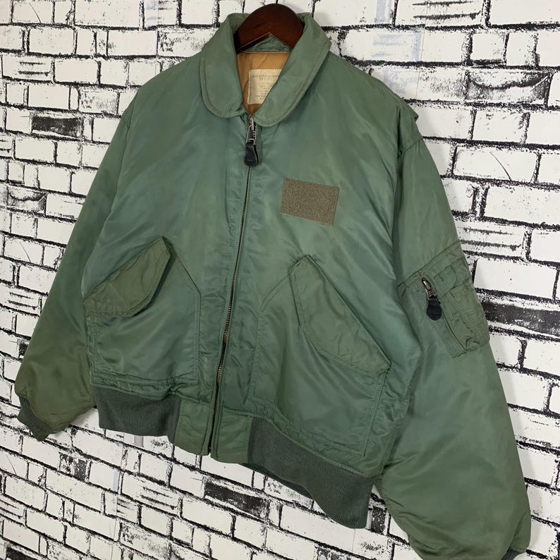 May include: A green bomber jacket with a zipper closure and a patch on the left side of the chest. The jacket has a ribbed collar, cuffs, and waistband.